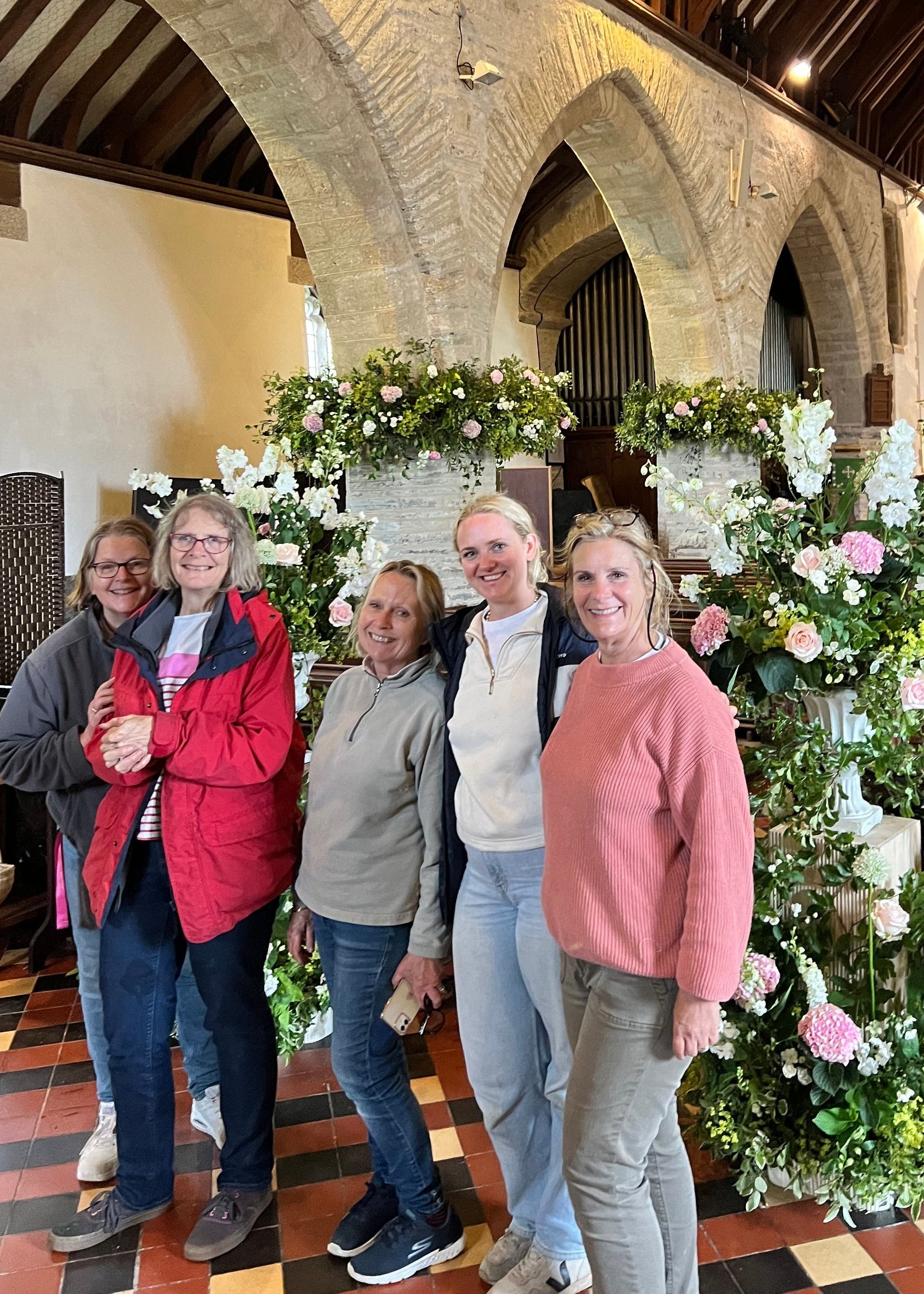 Five women posing together inside a historic church hall, standing in front of a large floral arrangement with pink and white flowers and green foliage.