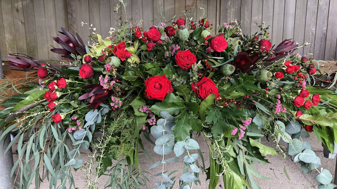 A large floral arrangement featuring vibrant red roses, deep burgundy foliage, green eucalyptus, and pink filler flowers, displayed outdoors against a wooden fence.