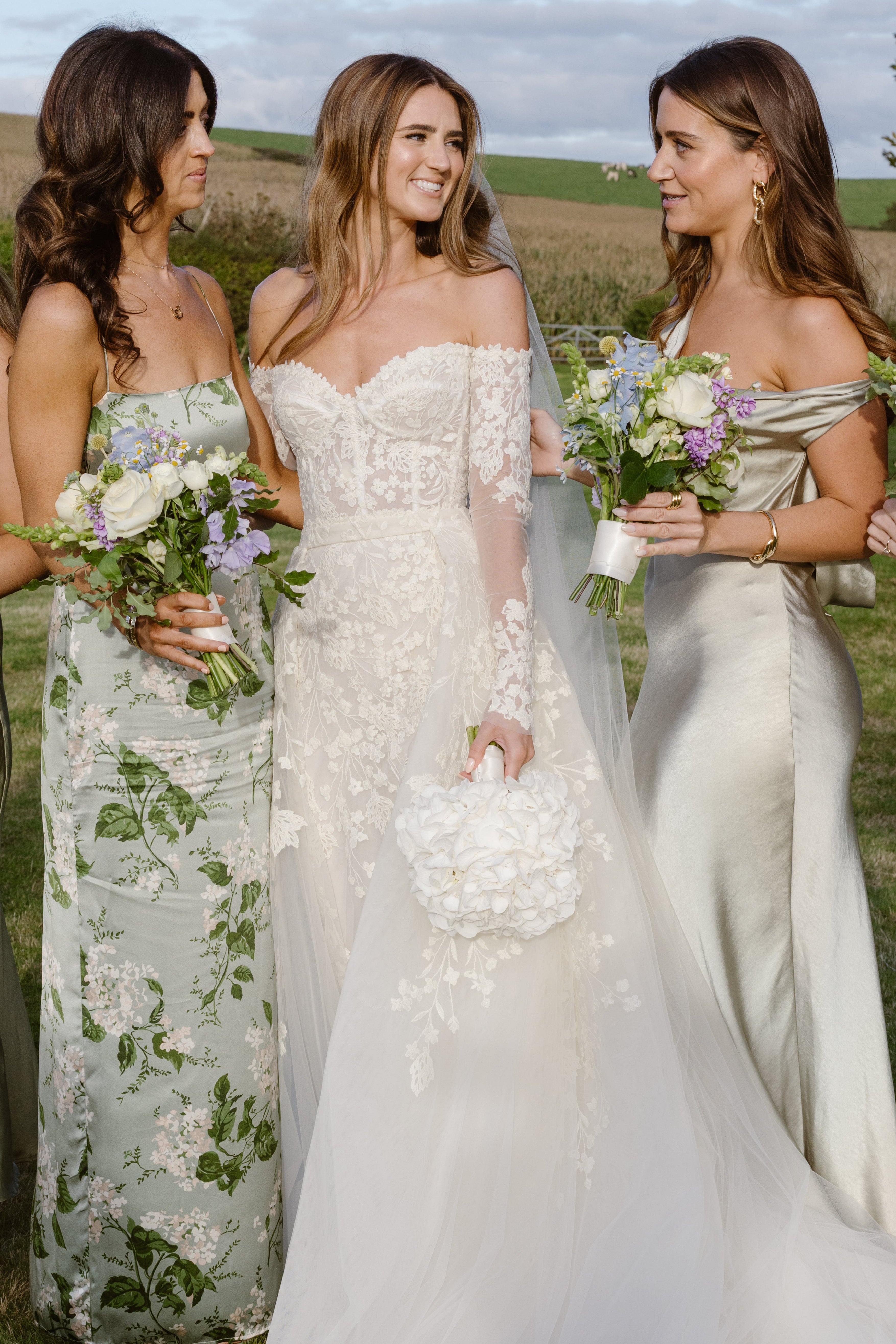A bride in an off-the-shoulder lace wedding gown stands outdoors with bridesmaids in floral and satin dresses, holding bouquets in a grassy field under a cloudy sky.