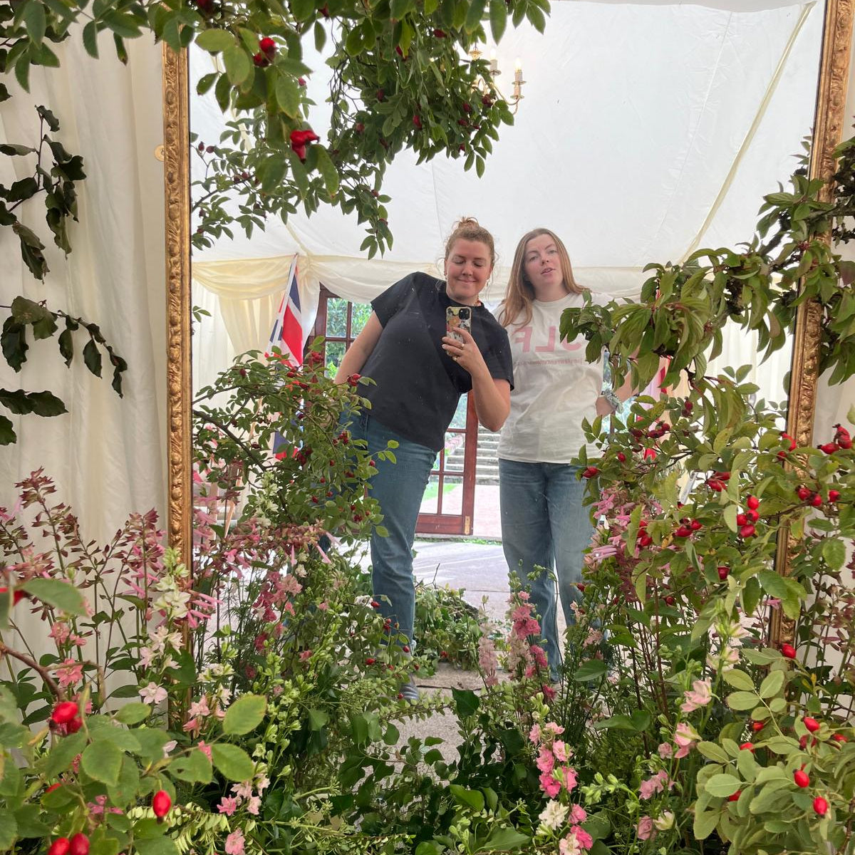 Two women posing for a mirror selfie inside a large, ornate gold-framed mirror surrounded by lush green rose bushes with red blossoms under a white canopy tent.