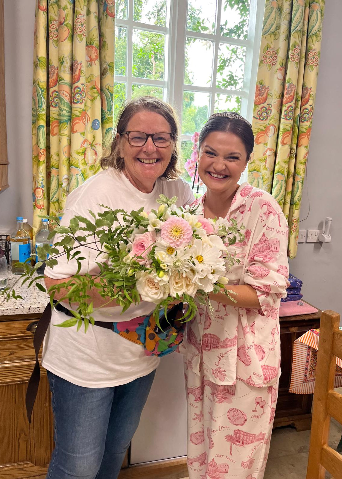 Two women smiling in a kitchen setting, holding a large bouquet of pink, white, and green flowers. One woman wears a white t-shirt and jeans, and the other wears a pink flamingo-patterned pajama set.